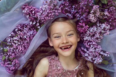 Child girl in a purple floral dress lies on the ground among lilacs on a veil in spring