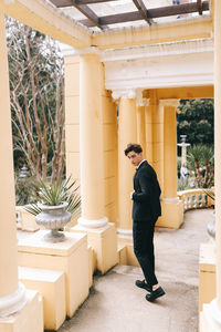 A beautiful young man, the groom in an elegant wedding suit, stands posing in the city's old park