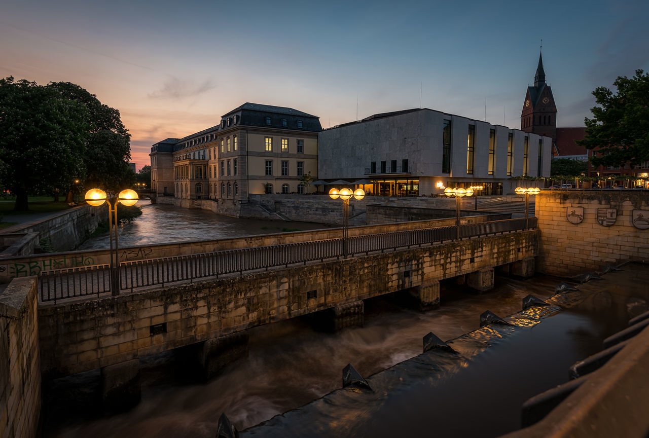 ILLUMINATED BRIDGE OVER RIVER BY BUILDINGS IN CITY AGAINST SKY