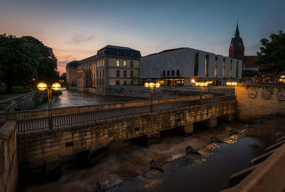 Illuminated bridge over river by buildings in city against sky at dusk