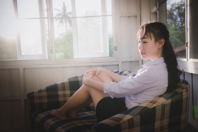 Side view of young woman sitting on chair against window