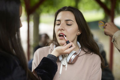 Teenage girl applying lipstick to female friend at park