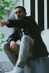 Low angle view of young man pointing while sitting on seat against wall