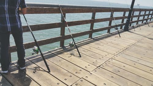 Low section of man standing on pier by fishing poles at railing against sea