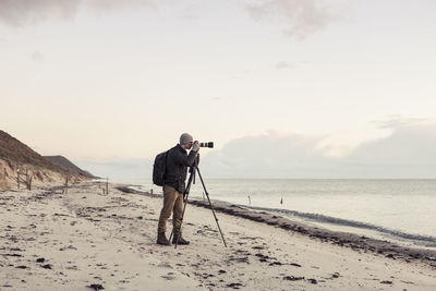 Full length side view of hiker photographing through slr camera on shore at beach