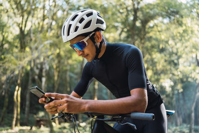 Male biker in helmet and cycling glasses text messaging on cellphone while sitting on bicycle in woods on sunny day