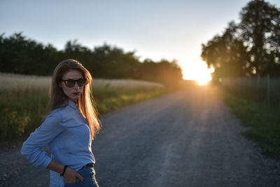 Portrait of young woman standing by road against sky during sunset