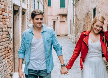 Smiling young couple holding hands while walking on footpath against buildings in city