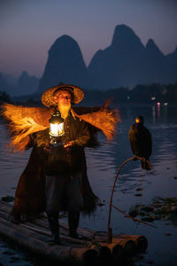 Man with lantern standing by bird on boat in lake against sky during sunset