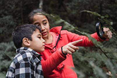 Ethnic child with magnifying glass demonstrating fern plant to sibling while exploring forest in daytime
