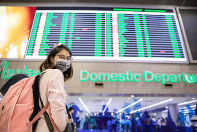 Woman tourist wearing mask standing with pink backpack and looking time board at airport