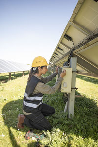 Side view of female engineer repairing fuse box while kneeling near solar panels at power station