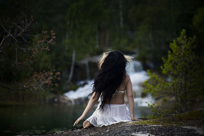 Rear view of woman sitting against waterfall in forest