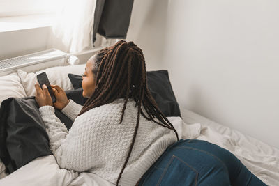 Rear view of woman using mobile phone while lying on bed at home