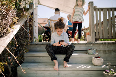 Girl using mobile phone while sitting on steps with sibling in background at patio
