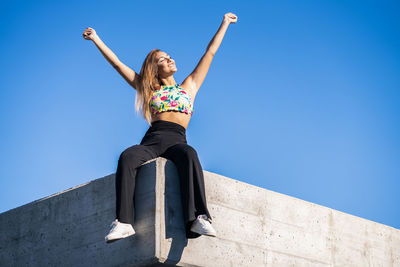 Low angle view of woman with arms raised sitting on retaining wall against clear blue sky