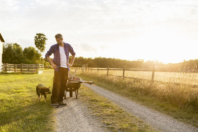 Man with dog and wheelbarrow standing on dirt road against clear sky