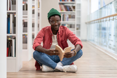 Happy african american millennial guy sitting on floor reading book while visiting library