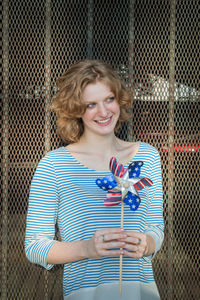 Happy woman looking away while holding pinwheel toy against fence