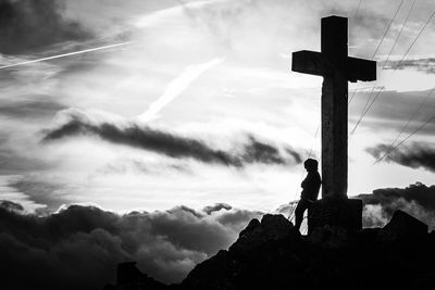 Silhouette young man standing by cross against cloudy sky during sunset