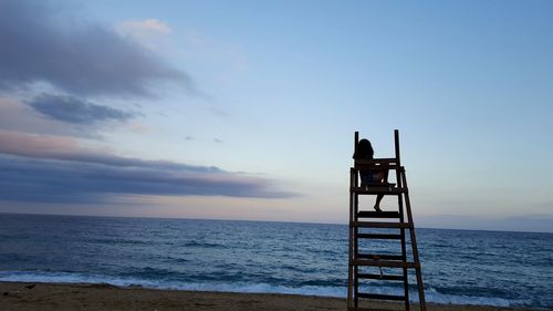 Woman sitting on wooden chair at beach against sky