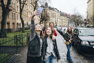 Smiling girl taking selfie through smart phone with friends while standing on sidewalk in city