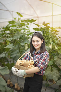 Portrait of smiling young woman carrying vegetables while standing against plants in greenhouse