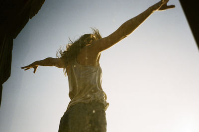 Low angle view of woman standing with arms outstretched against clear sky seen through glass