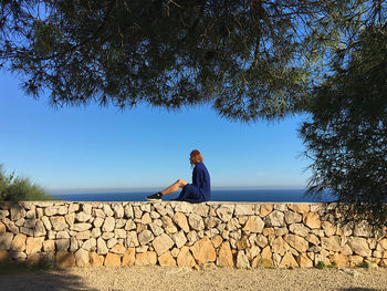 Man sitting on retaining wall against blue sky