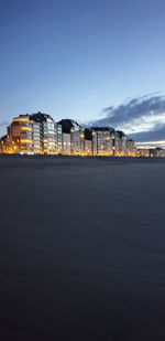 Beach with illuminated buildings in background against blue sky during sunset