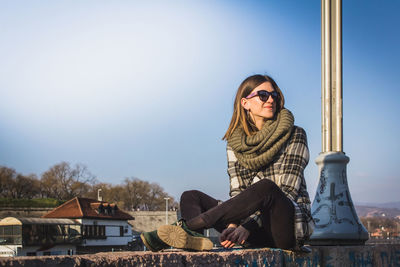 Portrait of smiling young woman sitting against built structure against clear sky