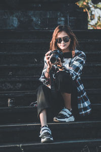 Full length of young woman photographer wearing sunglasses is sitting outdoors