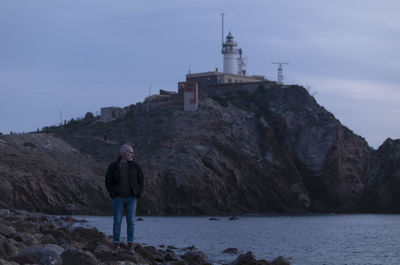 Adult man in black jacket standing on rocky beach looking at sea with lighthouse. almeria, spain