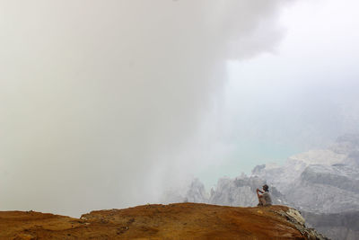 Man looking at fog while sitting on mountain against sky