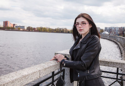 Young woman standing on railing by river against sky in city