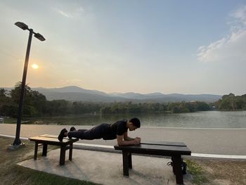 Man relaxing on bench by lake against sky during sunset