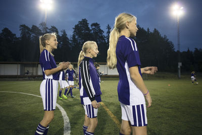 Side view of girls standing on soccer field against sky at dusk