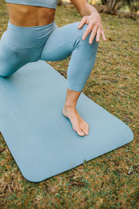 Crop unrecognizable barefoot slim female in blue leggings stretching body in crescent lunge on the knee pose while practicing yoga in park