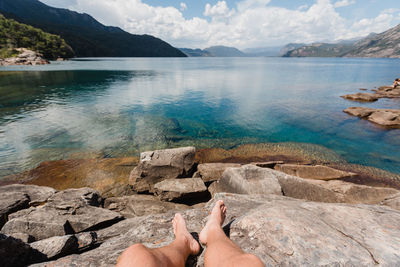 Low section of person on rock by lake against sky