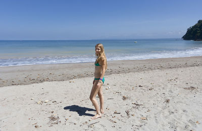 Young woman wearing bikini standing on sea shore at beach against blue sky