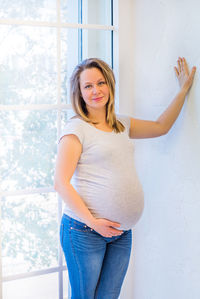 Portrait of pregnant woman standing against window at home