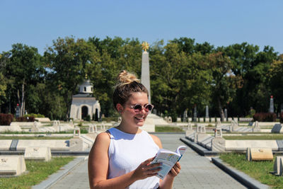 Smiling woman in sunglasses reading book against trees