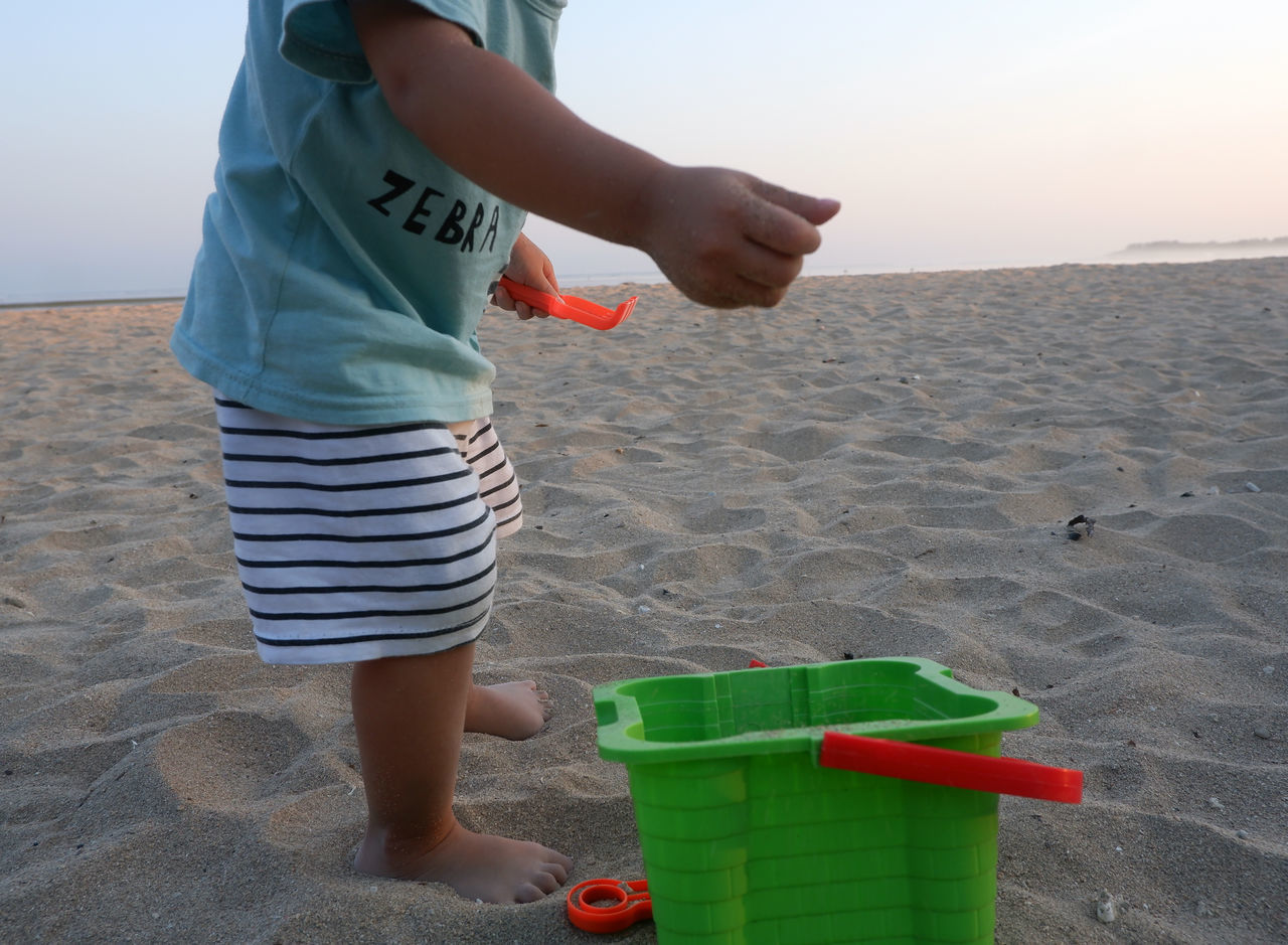 beach, land, one person, sand, childhood, nature, standing, child, water, sea, container, sky, casual clothing, leisure activity, bucket, day, holiday, women, vacation, environment, trip, lifestyles, holding, men, outdoors, barefoot, female, adult, summer, person, sunlight, full length, sandal, clear sky