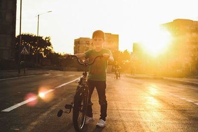 Portrait of boy with bicycle standing on road against sky during sunset