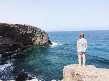 Rear view of young woman standing on cliff by sea against clear sky