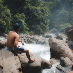 Shirtless man sitting on rock by stream in forest