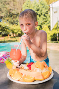 Cute little shirtless boy with wet blond hair drinking glass of fresh juice with straw and looking at camera after swimming in outdoor pool on sunny summer day