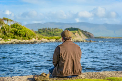 Rear view of man sitting by sea against sky