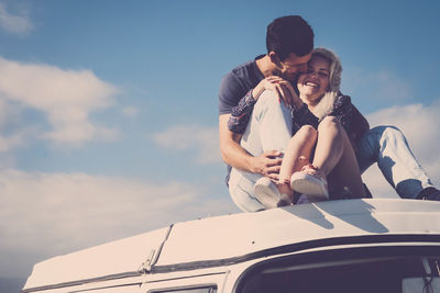 Young couple sitting on car against sky