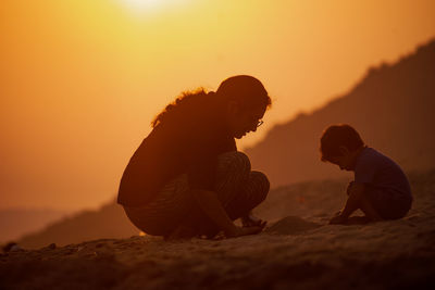 Rear view of man sitting on rock against sky during sunset